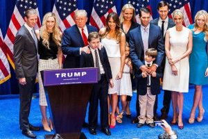 Familia Trump posando en un evento con banderas de fondo y un letrero de Trump en el frente.